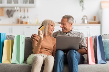 Neşeli bir çift rahat bir kanepede otururken dizüstü bilgisayarla online alışveriş yapmaktan zevk alır. Yakınlarda renkli alışveriş çantaları düzenlenir, canlı bir atmosfer yaratır..