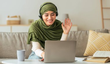 Yeşil eşarp takan genç bir kadın kulaklık takıyor ve dizüstü bilgisayarına el sallarken gülümsüyor. Oturma odasındaki kanepede oturup video görüşmesinin tadını çıkarıyor. Yakınlarda bir kupa ve kitap var..