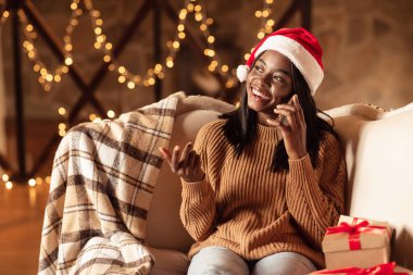 Noel şapkası takan bir kadın rahat bir koltukta oturur, gülümser ve telefonda sohbet eder. Şenlikli süslemeler ve paketlenmiş hediyeler sıcak aydınlık odadaki tatil ruhunu artırıyor..