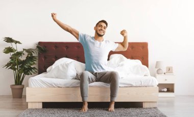 Genç bir adam yatağının kenarında oturur, gülümserken kollarını uzatır. Güneş ışığı odaya girer ve neşeli bir sabah atmosferi yaratır. Bir bitki doğaya bir dokunuş ekler..