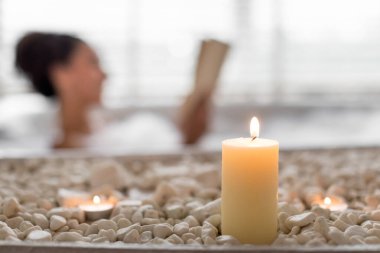 Bir kadın, sakin bir spa ortamında huzurlu bir akşam geçirir. Etrafı mumlar ve pürüzsüz çakıl taşlarıyla çevrili, sakin bir atmosferde kitap okurken..