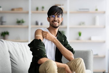 Genç bir adam evdeki bir kanepede rahatça oturur, aşıyı aldıktan sonra bol bol gülümser. Kolundaki bandajı gösteriyor ve sağlığa hazır olduğunu vurguluyor..