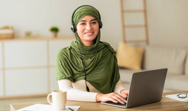 Yeşil başörtüsü takmış bir kadın ahşap bir masada oturuyor, bir fincan kahvenin keyfini çıkarırken dizüstü bilgisayarına odaklanıyor. Konforlu ev ortamında bitkiler ve yumuşak mobilyalar yer alıyor..