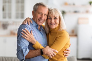 Yaşlı bir çift rahat bir yuva ortamında birbirlerine sarılırken sıcak bir şekilde gülümsüyor. Doğal ışıkla dolu güzel dekore edilmiş bir salonda şefkat gösteriyorlar..