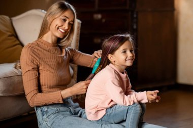 Bir anne kızının saçını nazikçe tararken gülümser. Onlar iyi ışıklandırılmış bir oturma odasında, birlikte sıcak ve eğlenceli bir anın tadını çıkarıyorlar..