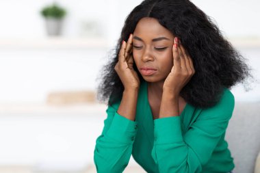 Yeşil kazaklı bir kadın gözlerini kapar ve stres ya da yorgunluk belirtileri göstererek şakaklarını tutar. Tarafsız bir arka planı olan kapalı bir ortamda oturuyor..