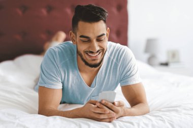A young man relaxes on his stomach on a bed, smiling as he checks his smartphone. The setting is cozy with soft white bedding and a dark headboard.