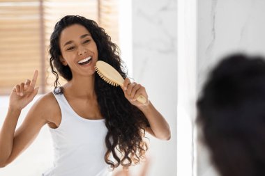Uzun kıvırcık saçlı bir kadın, parlak ve çağdaş bir banyoda saçını tararken gülümsüyor. Aynadaki yansımasından zevk alıyor gibi görünüyor..
