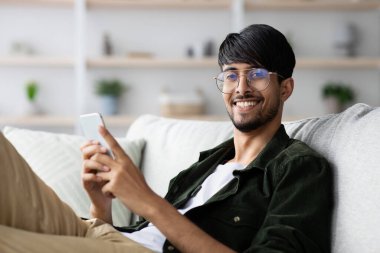 Gözlüklü genç bir adam iyi dekore edilmiş bir oturma odasındaki bir kanepede rahatça oturur. Akıllı bir telefon tutarken gülümsüyor ve boş zamanlarının tadını çıkarıyor..