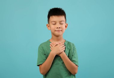 Genç bir çocuk ellerini göğsüne koyup bir anlık minnettarlığın tadını çıkarırken gözleri kapalı ve sakin bir ifadeyle ayakta duruyor. Arka plan yumuşak mavi bir renktir..