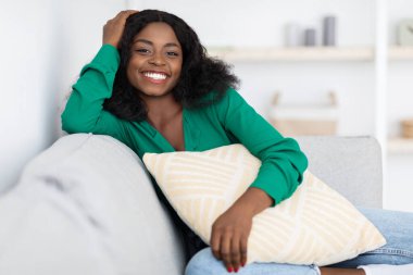 Kıvırcık saçlı bir kadın açık gri bir kanepede rahatça otururken gülümsüyor. Dekoratif bir yastık tutuyor ve yaşam alanının rahat atmosferini seviyor..