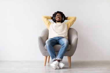 Genç siyahi bir adam, yumuşak bir koltukta oturup rahatlamanın tadını çıkarır. Kablosuz kulaklık takıyor ve müzik dinlerken gülümsüyor. Huzurlu bir atmosfer yaratıyor..