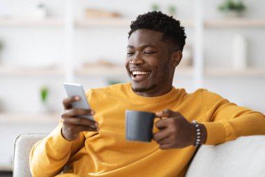 Mutlu Afro-Amerikan milenyumu çocuğu evdeki kanepede rahat rahat yaslanıyor, kahve içiyor ve modern akıllı telefon kullanıyor, kız arkadaşıyla sohbet ediyor, internette sörf yapıyor, uzayı kopyalıyor.