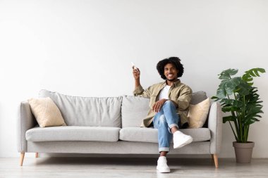 Genç siyah bir adam, klimayı ayarlamak için uzaktan kumanda kullanırken gülümseyerek rahat bir kanepede dinlenir. Sıradan giyinmiş ve etrafı rahat ev dekorlarıyla çevrili..