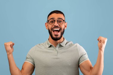 Gözlüklü neşeli bir adam zafer için yumruklarını kaldırarak heyecan ve neşe gösteriyor. Açık yeşil bir polo tişört giyiyor ve düz mavi bir arkaplanda duruyor..