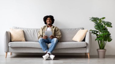 Genç bir Afrikalı Amerikalı adam modern bir kanepede rahatça oturuyor. Elinde dijital bir tablet tutarken gülümsüyor, boş zamanlarında bitkilerle dolu parlak bir oturma odasında eğleniyor..