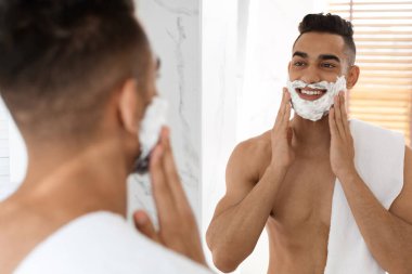 Genç bir adam iyi ışıklandırılmış bir banyoda duruyor, yüzünü tıraş ederken gülümsüyor. Yüzünde tıraş kremi var ve elinde jilet var. Bakım rutininden kendine güveni ve zevki yayıyor..