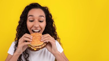 Kıvırcık saçlı bir kadın hamburgerden büyük bir ısırık almaya hazırlanırken heyecanlı görünür. Parlak sarı arka plan o ana enerji katar, onu canlı ve eğlenceli hissettirir..