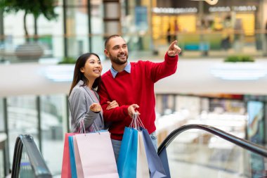 Genç bir çift ellerinde renkli alışveriş torbalarıyla bir alışveriş merkezinde yürüyen merdivene binerken gülümsüyor. Bulgularını heyecanla tartışarak neşeli bir anı paylaşıyorlar..