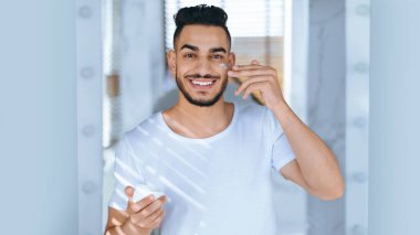 Genç bir adam iyi aydınlatılmış bir banyoda yüzüne krem sürerken gülümsüyor. Güneş ışığı pencereden içeri giriyor, tımar rutinini vurguluyor..