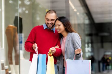 Mutlu çift şık bir perakende mağazasının önünde renkli alışveriş torbaları tutarken el ele yürür. Gülücüklerini paylaşıyorlar ve birlikte alışverişe çıkıyorlar..