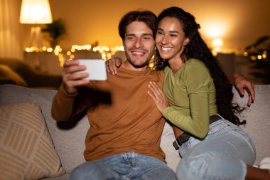 Genç bir çift, akıllı telefonlarıyla selfie çekerken sarılıp gülümsüyor. Evdeki rahat bir kanepede oturuyorlar. Birlikte rahat bir hafta sonunun tadını çıkarıyorlar..