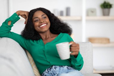 Gülümseyen genç bir kadın elinde bir bardakla koltukta oturuyor, rahat ve rahat görünüyor oturma odasında, yumuşak dekor ve doğal ışıkla dolu..