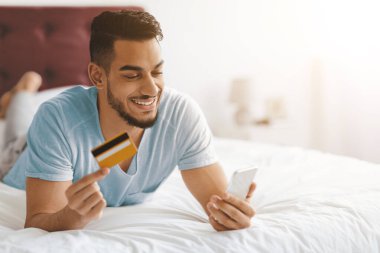Bir erkek yatakta yatarken online alışveriş yapmaktan mutlu olur. Bir elinde kredi kartı tutuyor ve akıllı telefonuna gülümseyerek bakıyor. Odanın yumuşak ışıklandırması ve rahat bir atmosferi var..