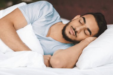 Sakallı genç bir adam yatağında uzanıyor, derin uykuda. Açık mavi bir gömlek giyiyor ve sakin bir atmosferde yumuşak beyaz battaniyelerle çevrili sakin bir anın tadını çıkarıyor..