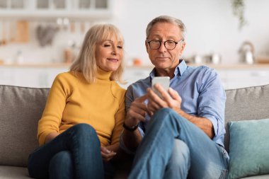 Yaşlı bir çift rahat bir kanepede oturur, akıllı telefona bakarken bir anı paylaşırlar. Arka planda yumuşak ışıklandırma ve modern bir mutfak görülebilir..