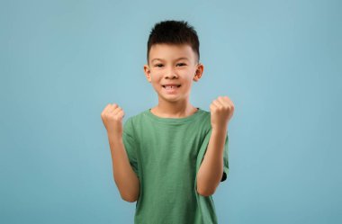 Kısa siyah saçlı neşeli genç bir çocuk açık mavi bir fon karşısında yumruklarını kaldırıyor. Neşeli ifadesi bir zafer ve mutluluk anı sergiliyor..