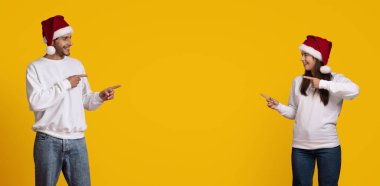 Noel şapkası takan iki arkadaş gülüyor ve neşe içinde birbirlerini gösteriyorlar. Samimi beyaz süveterler giyiyorlar, canlı sarı bir arka plana karşı, somut bir tatil ruhu..