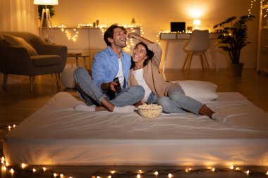 Neşeli çift loş bir odada gevşiyor, televizyon izlerken gülüyor ve birbirlerine patlamış mısır yediriyorlar. Sıcak atmosfer mükemmel bir hafta sonu eğlencesi anı yaratır..