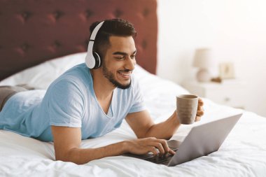 Genç bir adam önünde dizüstü bilgisayarla yatar. O, kahve yudumlarken kulaklık takıyor ve gülümsüyor. Odanın sakin bir atmosferi var. Yumuşak ışıklandırma ve rahat bir yatak..