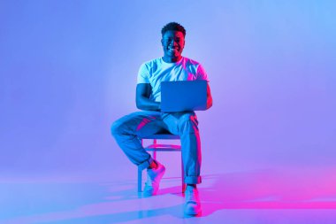 Genç bir adam bir sandalyede oturup dizüstü bilgisayarla çalışırken gülümsüyor. Sahne parlak mavi ve pembe ışıklarla aydınlatılıyor, modern ve neşeli bir atmosfer yaratıyor..