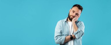 Sakallı ve gözlüklü genç bir adam parlak mavi bir arka plana karşı duruyor. Yüzünde eli var ve gündelik kıyafetlerle dalgın dalgın dalgın dalgın dalgın dalgın dalgın dolaşıyor..