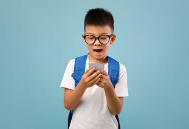 Gözlüklü ve beyaz tişörtlü genç bir çocuk akıllı telefonuna bakarken heyecanını ifade ediyor. Üzerinde mavi bir sırt çantası var, canlı bir atmosfer yaratıyor..