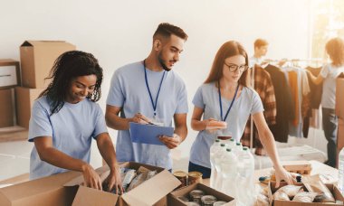 Üç gönüllü, bir sosyal merkezde yiyecek ve temel şeyleri kutulara ayırıyor. Bağış organize etmekle meşgulken, diğer insanlar arka planda kıyafet hazırlıyorlar..