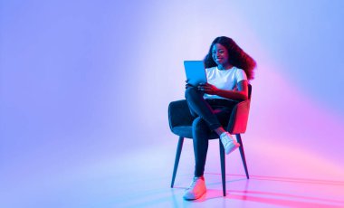 Kıvırcık saçlı genç bir kadın tabletiyle meşgul olan bir sandalyeye rahatça oturur. O gülümserken, hareketli arka plan hareketli bir atmosfer yaratıyor ve hareketlerinin tadını çıkarıyor..