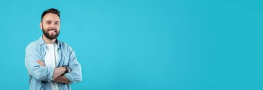 Sakallı genç bir adam kolları çapraz dururken kendine güvenerek gülümsüyor. Sıradan bir kıyafet giyiyor ve canlı, mavi bir arka plana yaslanıp neşeli bir atmosfer yaratıyor..