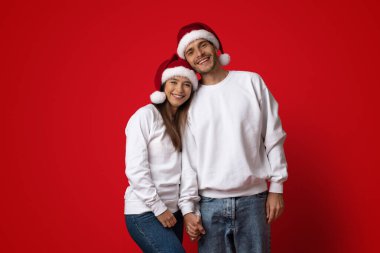 Her ikisi de Noel Baba şapkası takan neşeli bir çift el ele tutuşuyorlar. Sıradan beyaz eşofman üstü ve kot pantolon giyiyorlar, canlı kırmızı bir arka plana neşeyle gülümsüyorlar..