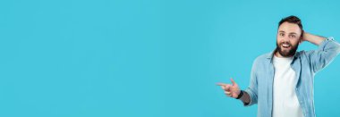 Sakallı bir adam parlak mavi bir arka plana karşı duruyor. Neşeli görünüyor ve bir gülümseme ile sağını işaret ediyor, muhtemelen ilginç ya da komik bir şey paylaşıyor..