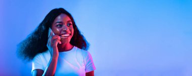 Kıvırcık saçlı neşeli genç bir bayan telefonda. Beyaz bir gömlek giyiyor ve renkli mavi ve pembe bir arka planda mutlu görünüyor, enerjik bir atmosfer yaratıyor..