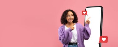 Genç bir kadın boş bir telefon ekranının yanında duruyor, gülümsüyor ve aşk tepkisi ikonlarına işaret ediyor. Arka plan yumuşak pembe, sosyal medya temalı kolaj için neşeli bir atmosfer yaratıyor..