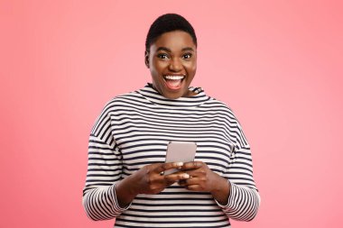 Neşeli bir kadın elinde akıllı bir telefonla pembe bir zemin üzerinde duruyor. Geniş bir gülümseme sergiliyor, mutlu bir an sergiliyor, muhtemelen internetten biriyle iletişim kuruyor..