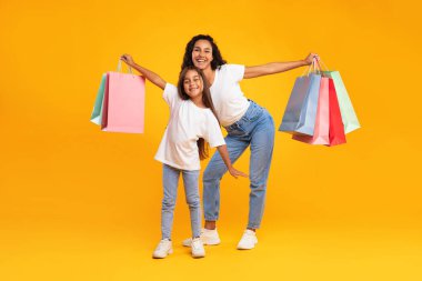 Neşeli bir anne ve genç kızı ellerinde renkli alışveriş torbalarıyla gülümsüyor. Parlak sarı bir arkaplanın önünde neşeli bir şekilde poz veriyorlar, eğlenceli günlerini sergiliyorlar.