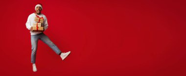 Noel Baba şapkası takan bir adam elinde hediyeler tutarken havaya sevinçle zıplıyor. Canlı kırmızı arka plan şenlik havasını artırır. Tatil ruhunun canlı bir kutlaması..