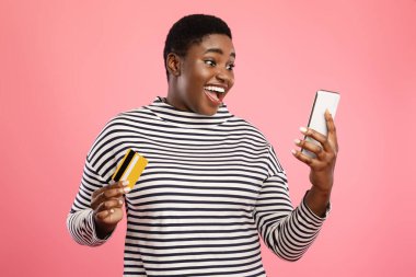 Neşeli bir birey, elinde bir kredi kartı ve akıllı bir telefon tutarken ışıl ışıl gülümser. Dekorun neşeli pembe bir arka planı var, enerjik bir ortam yaratıyor..