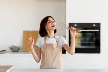 Genç bir kadın, parlak ve modern bir mutfakta çırpıcıyla şarkı söylemekten zevk alır. Günlük kıyafet ve önlük giyer, yemek hazırlarken mutluluk saçar..