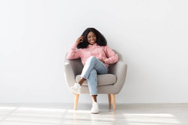 Pembe kapüşonlu neşeli genç bir kadın rahat gri bir sandalyede rahatlıyor. Gülümser ve elini çenesine koyar. Basit, şık bir odada doğal ışığın tadını çıkarır..